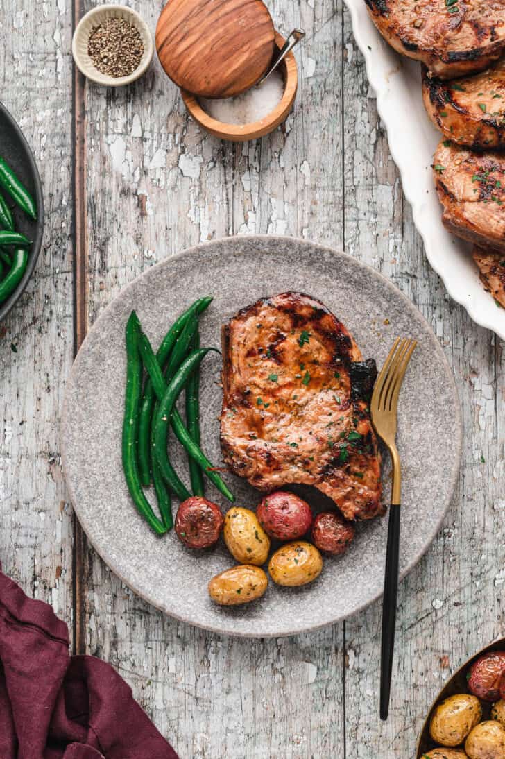 A grilled pork chop on a textured gray plate alongside roasted baby potatoes and green beans.