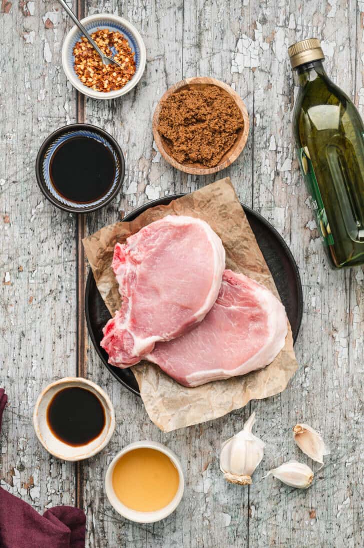 Ingredients for a grilled pork chop marinade laid out on a rustic gray surface, including brown sugar, red pepper flakes, oil, soy sauce and garlic.