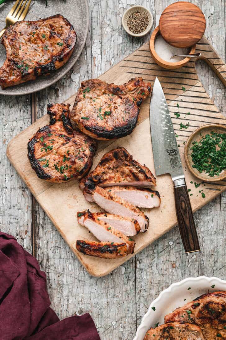 Grilled pork chops on a wooden cutting board with a large knife. One chop is sliced and the others are whole.