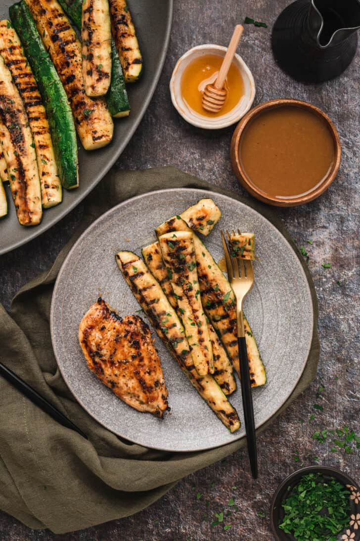 A gray plate with a piece of barbecue chicken breast and four spears of grilled zucchini, with a fork cutting the zucchini.