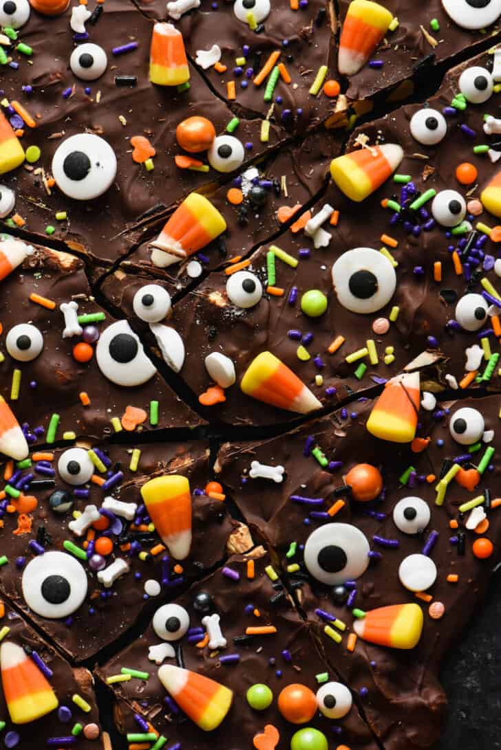 Halloween bark made from chocolate, sprinkles, candy corn and candy eyeballs, broken into pieces, on dark surface.