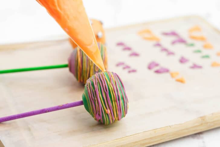 Candy coated donut holes on lollipop sticks being drizzled from an orange piping bag.