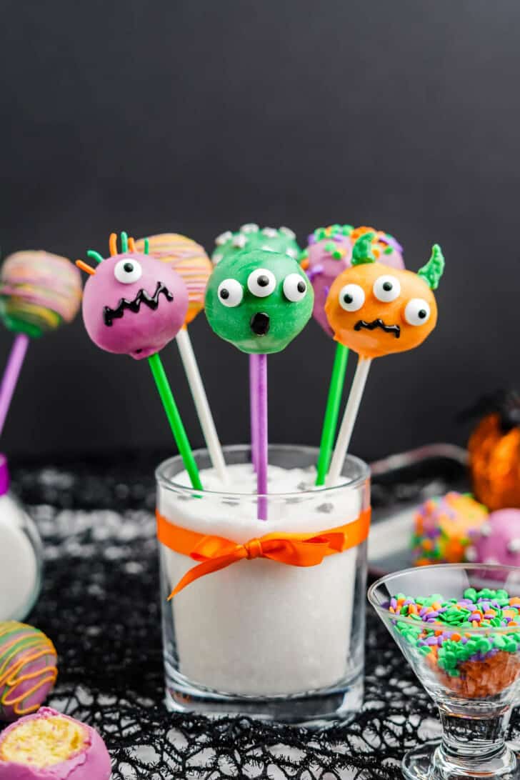 Purple, green and orange candy-coated Halloween cake pops decorated with candy eyeballs and sprinkles, sticking out a glass filled with white sugar and tied with an orange ribbon.