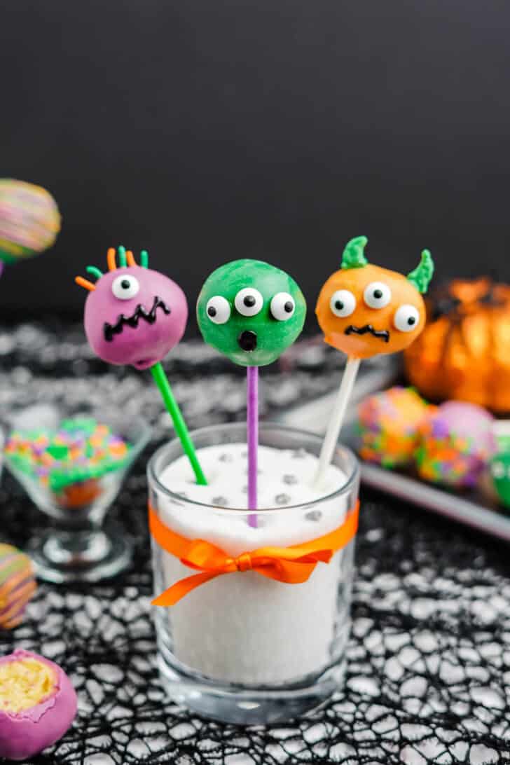 Purple, green and orange candy-coated Halloween cake pops decorated with candy eyeballs and sprinkles, sticking out a glass filled with white sugar and tied with an orange ribbon.