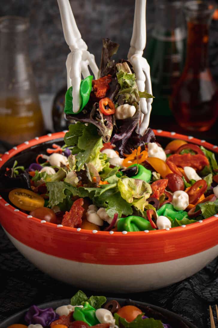 An orange bowl filled with with Halloween salad made with greens, olives, sliced peppers, dyed tortellini, mozzarella cheese skulls and pepperoni cut into bat shapes, with skeleton hands being used as salad tongs to lift salad out of the bowl from above.