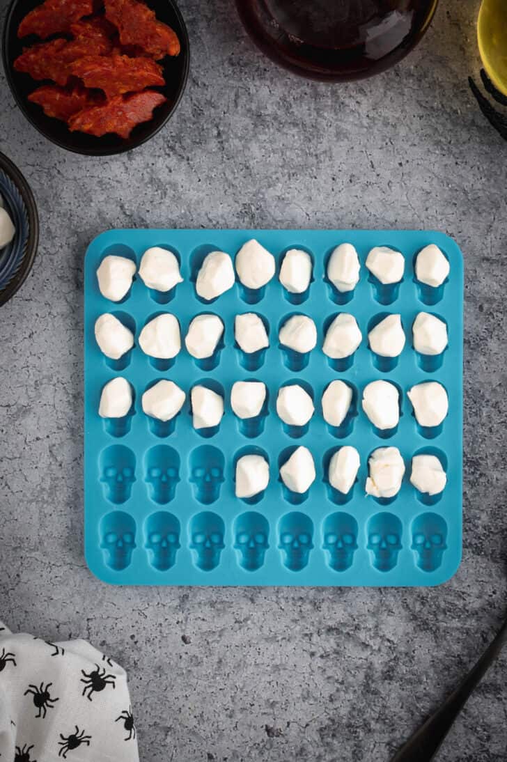 A blue silicone mold of skull shapes, with mozzarella cheese balls being placed into each skull form.