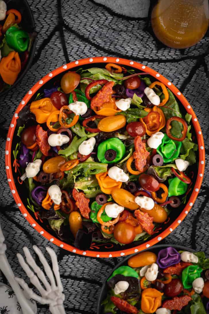 An orange bowl filled with with Halloween salad made with greens, olives, sliced peppers, dyed tortellini, mozzarella cheese skulls and pepperoni cut into bat shapes.