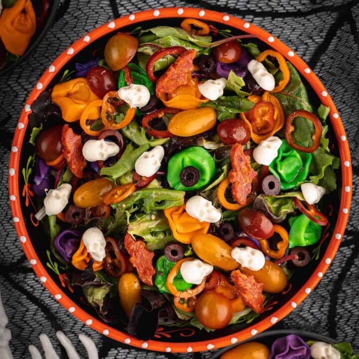 An orange bowl filled with with Halloween salad made with greens, olives, sliced peppers, dyed tortellini, mozzarella cheese skulls and pepperoni cut into bat shapes.