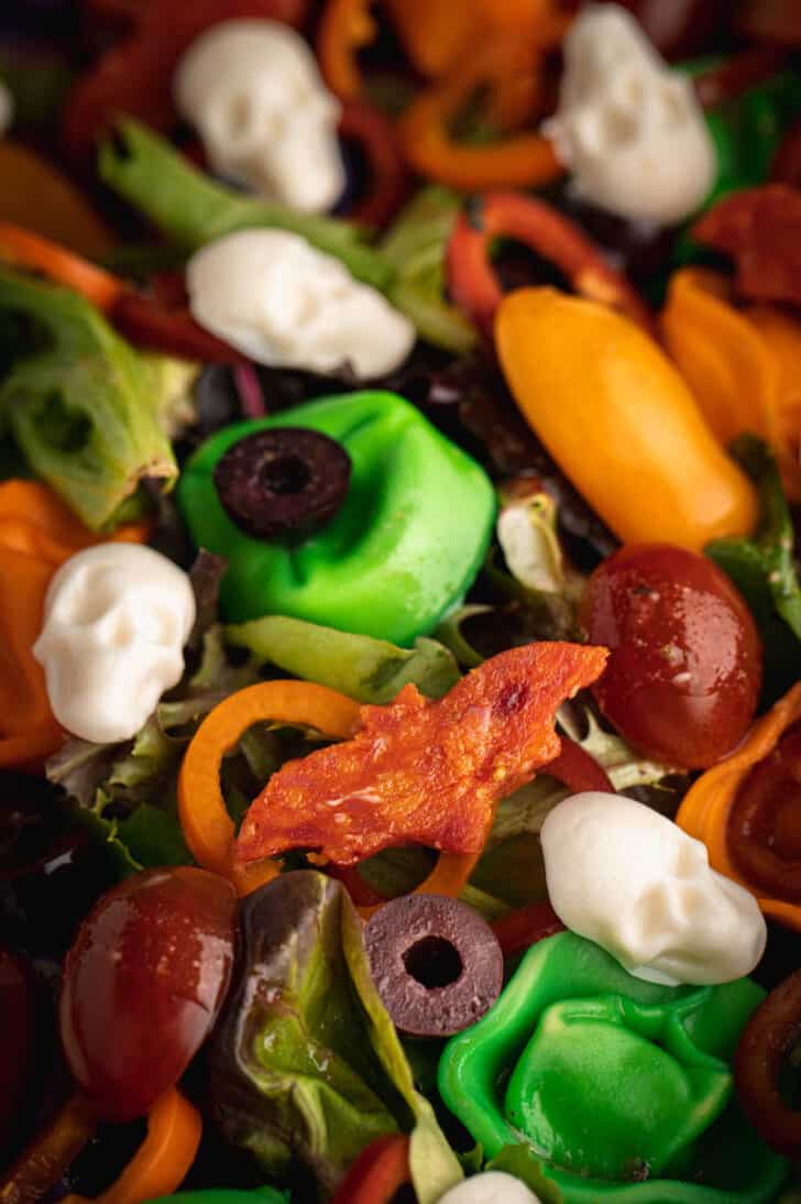 A Halloween side dish made with greens, peppers, tomatoes, olives, dyed tortellini, pepperoni cut into bat shapes and mozzarella cheese skulls.