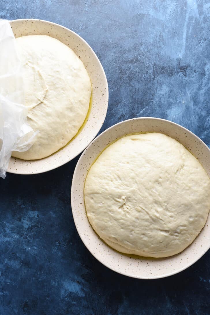 Two large speckled shallow bowls with risen homemade pizza dough in them.