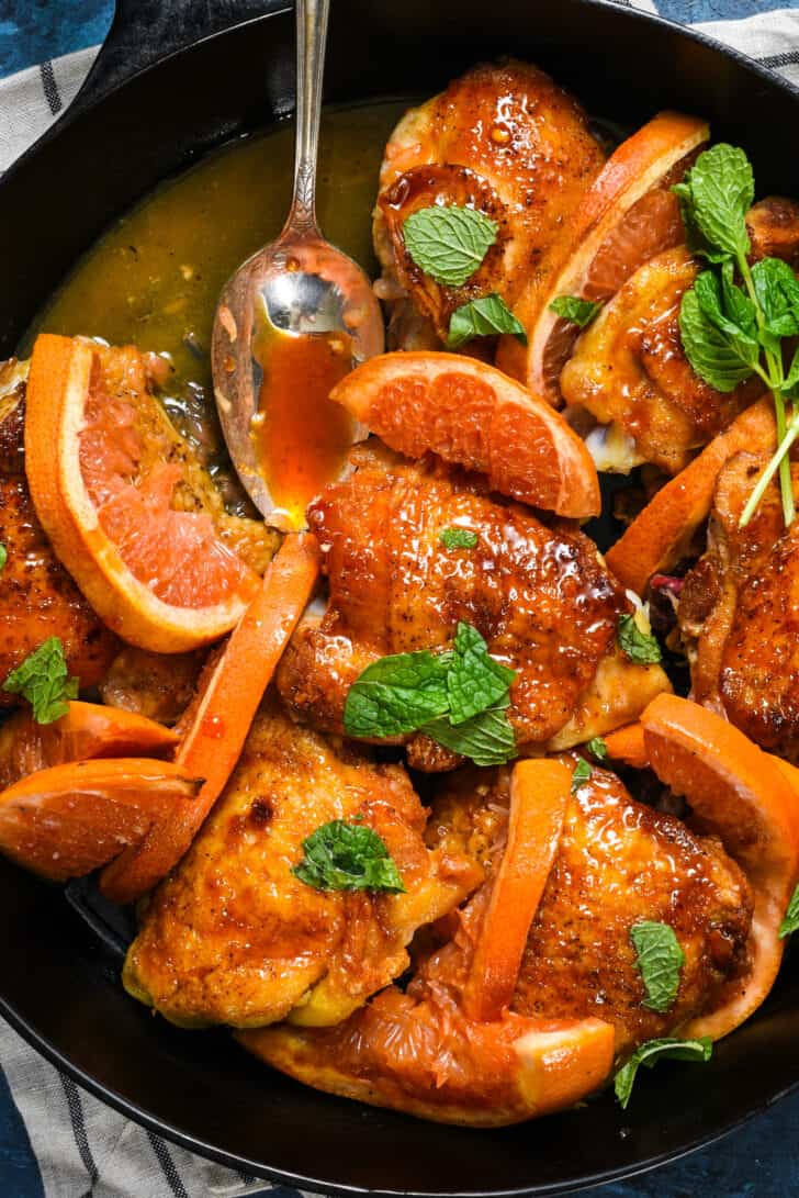 Cast iron skillet filled with honey orange chicken sprinkled with mint, with spoon scooping out sauce.