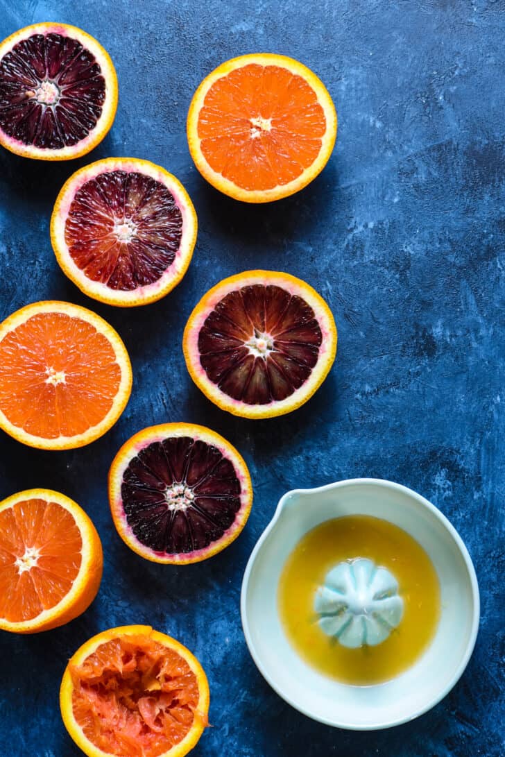 Halved blood oranges and Cara Cara oranges on blue background with light blue ceramic juicer.