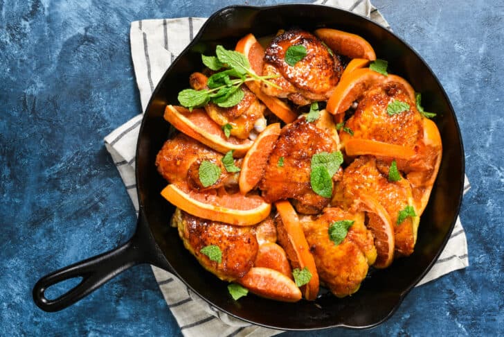 Cast iron skillet filled with chicken thighs topped with orange glaze and sprinkled with mint, with spoon scooping out sauce.