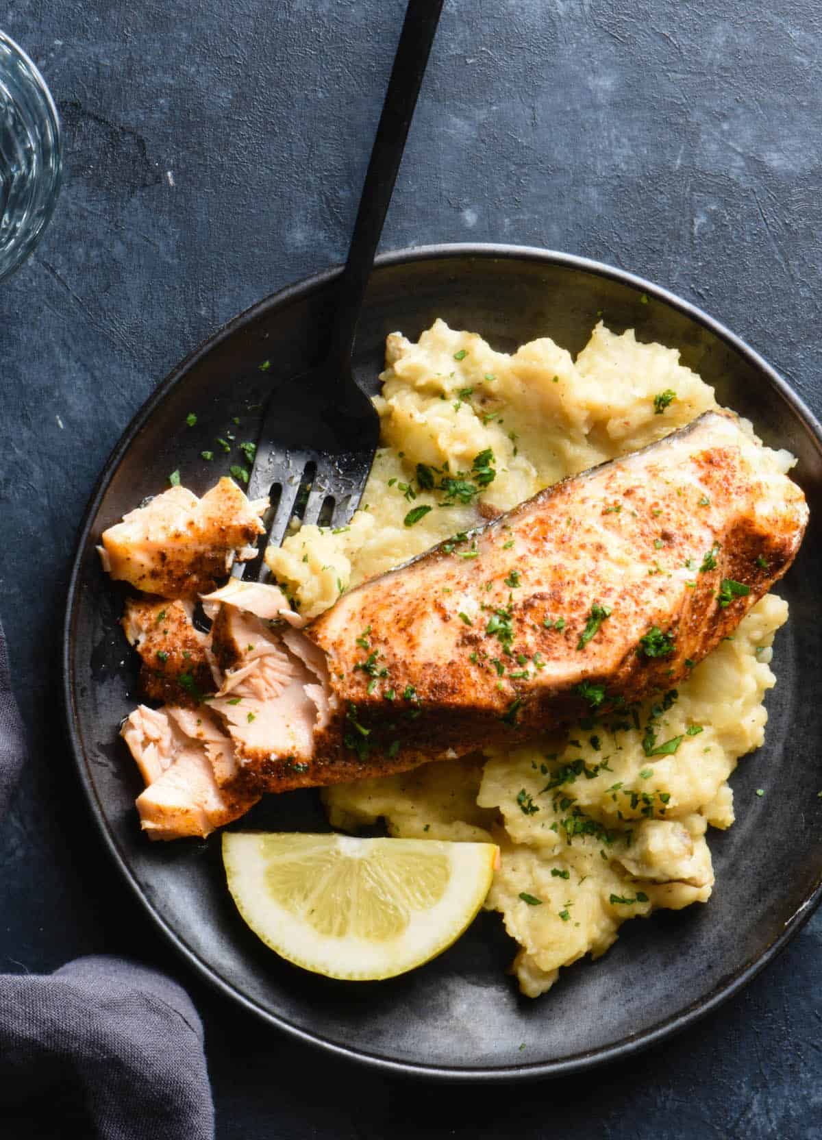 Black plate on dark background filled with mashed potatoes and salmon. Black fork is cutting into salmon fillet.