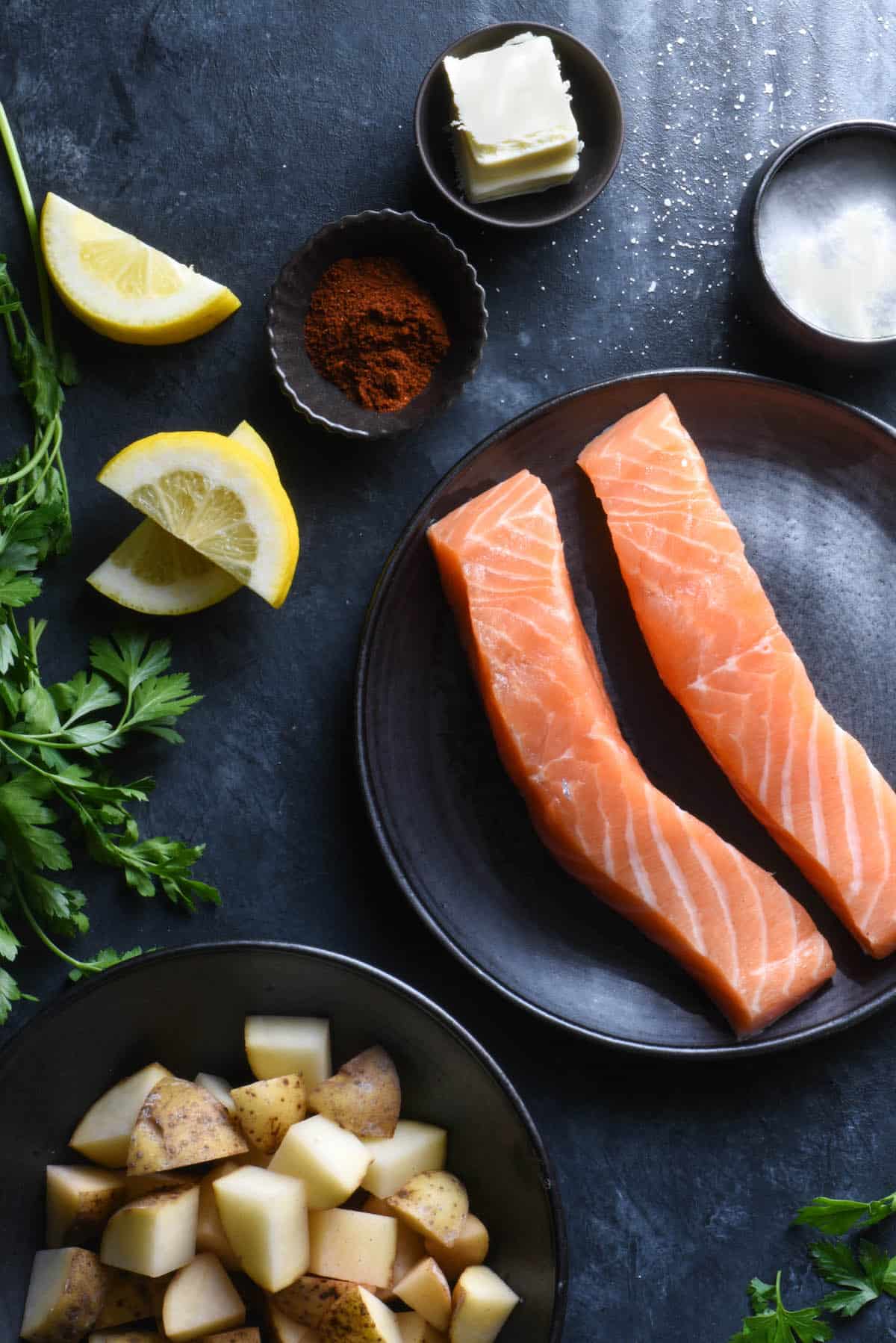 Dark background topped with ingredients: salmon fillets, raw chopped potatoes, parsley, lemon wedges, spices and butter.