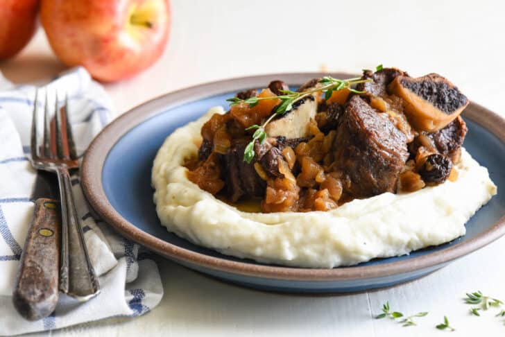 Shallow blue bowl filled with mashed potatoes, topped with Instant Pot beef ribs and garnished with thyme. Two apples are in background.