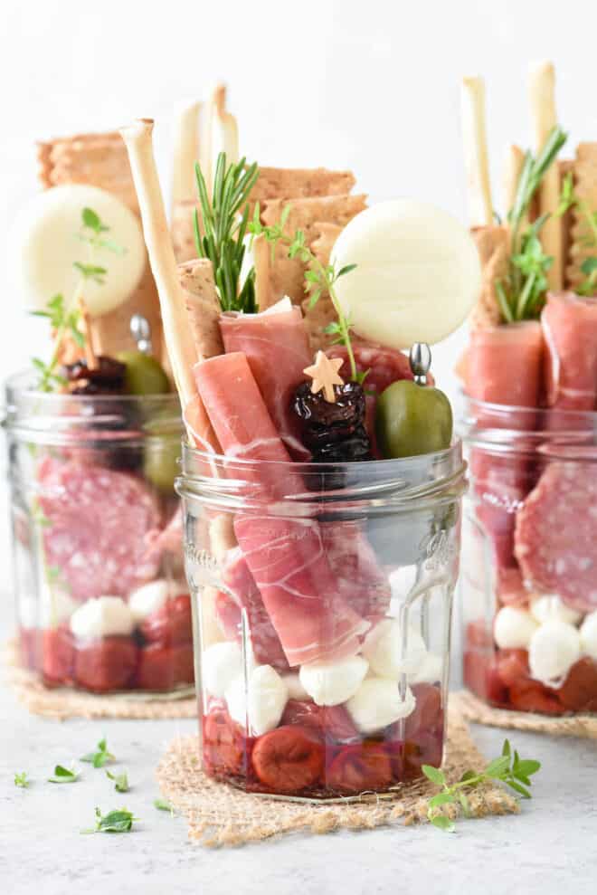 Three glass jars filled with cheese, fruit, cured meats, olives, crackers and herbs.