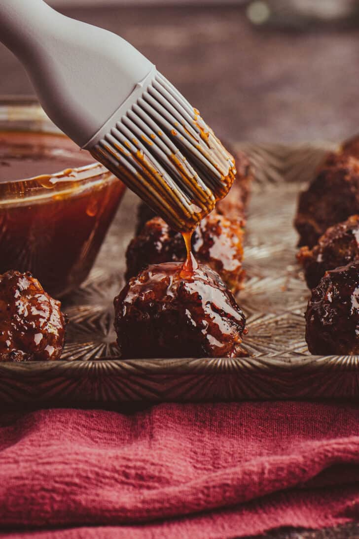 Gochujang meatballs on a textured baking pan being glazed with a deep red sauce by a gray silicone basting brush.