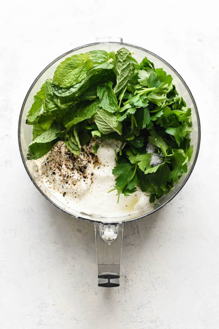 The ingredients for lamb lollipop sauce in the bowl of a food processor, including yogurt, mint, parsley, salt and pepper.