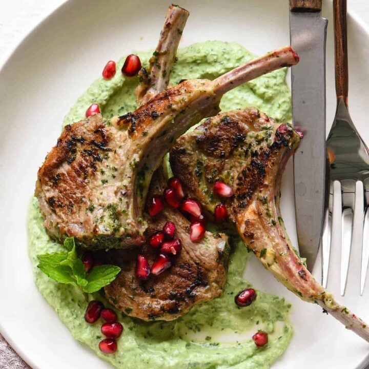 Lollipop lamb chops garnished with pomegranate seeds on top of a creamy green sauce, in a white shallow bowl.