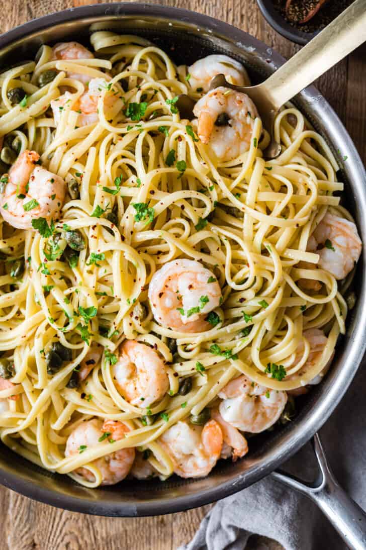 Stainless steel skillet filled with linguine and shrimp dressed with a caper and herb sauce.