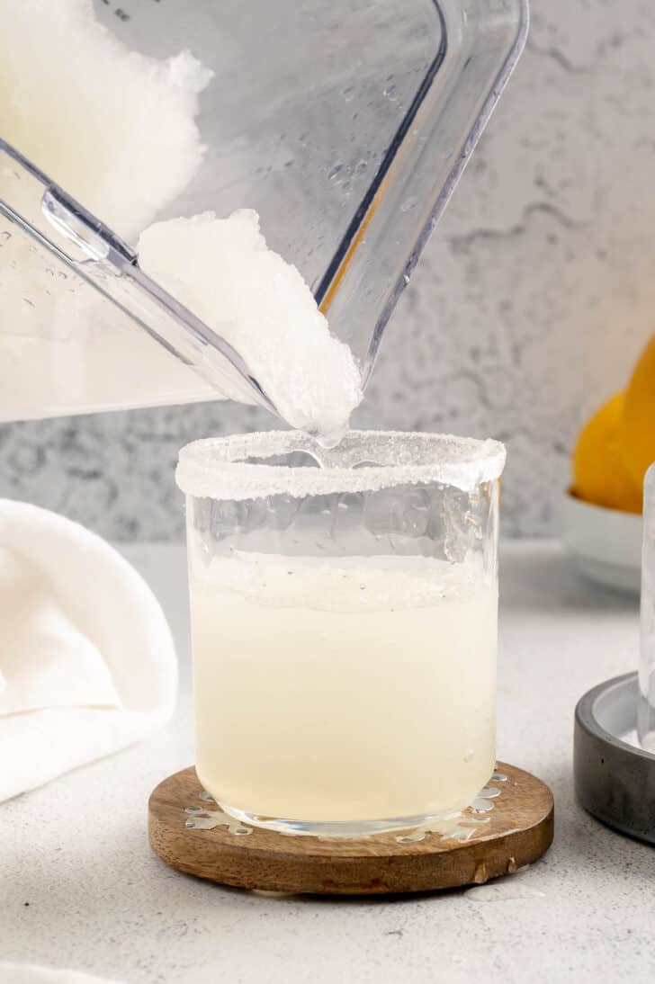 A clear blender pitcher pouring a mostly clear frozen liquid into a small glass on a wooden coaster.