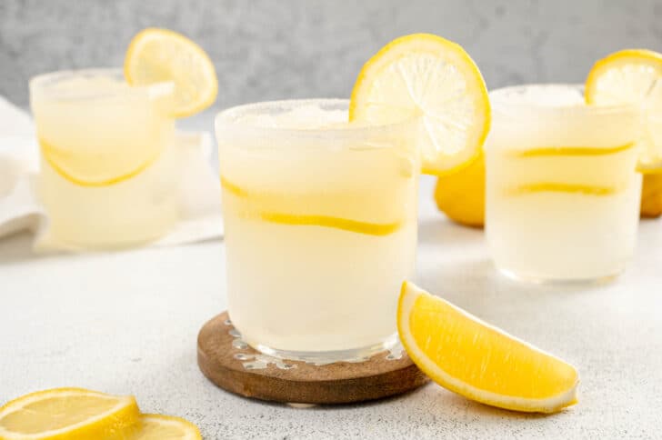 Tequila lemon cocktail in three glasses, garnished with lemon slices. The front glass is on a wooden coaster.