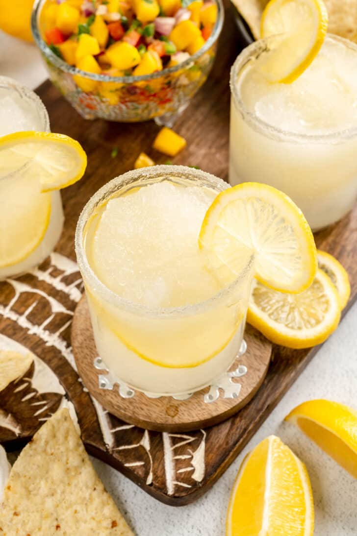 Lemon drop margaritas garnished with lemon slices, on a wooden serving board with mango salsa and tortilla chips.