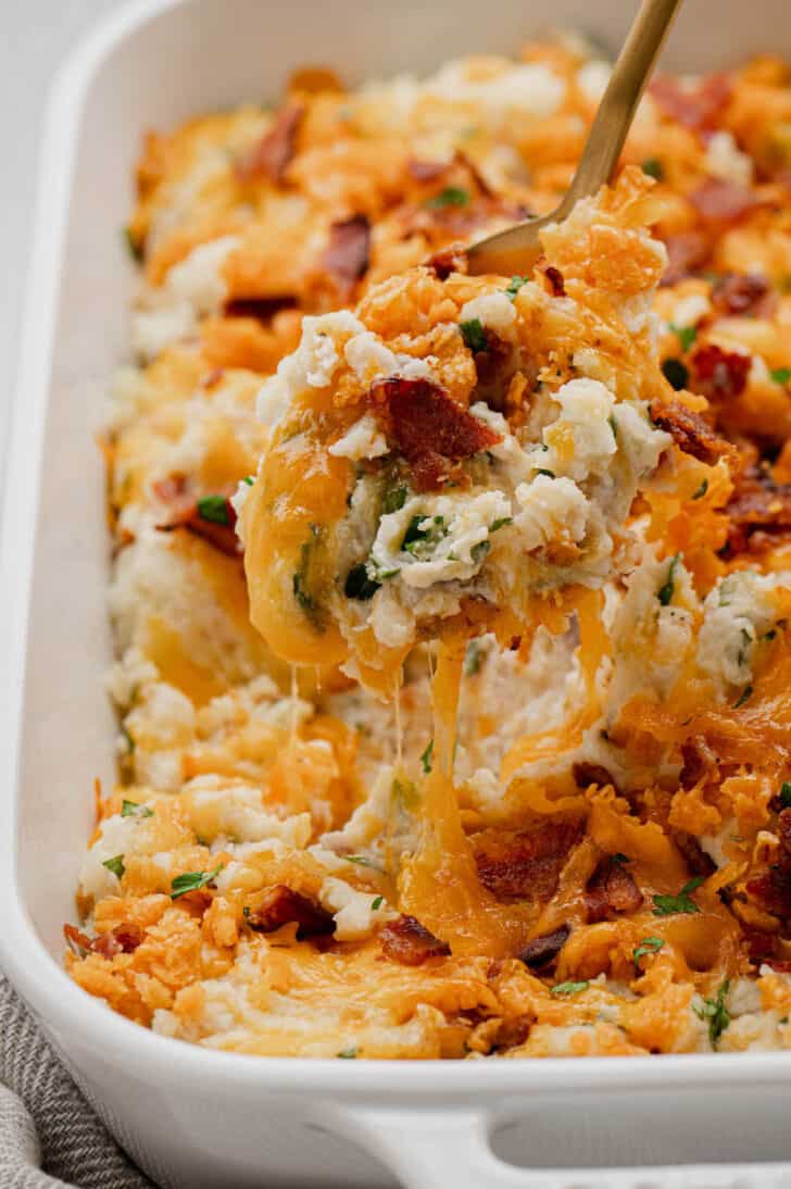 A gold spoon lifting a scoop of baked loaded mashed potatoes out of a baking dish.