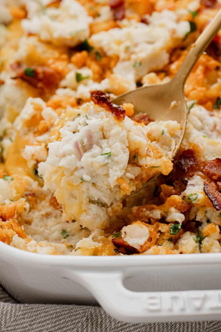 A gold spoon digging into loaded mashed potato casserole in a white baking dish.