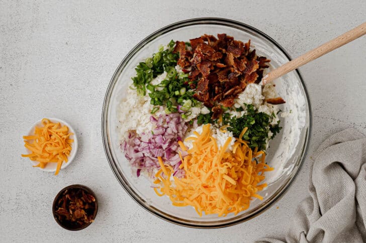 A glass mixing bowl filled with a creamy white mixture on the bottom, topped with bacon, onions, herbs, and shredded cheese.