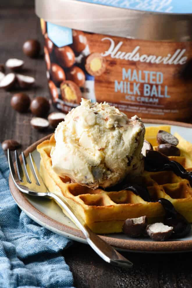 Waffle topped with hot fudge and ice cream on a plate, with a carton of Hudsonville Malted Milk Ball Ice Cream in the background.