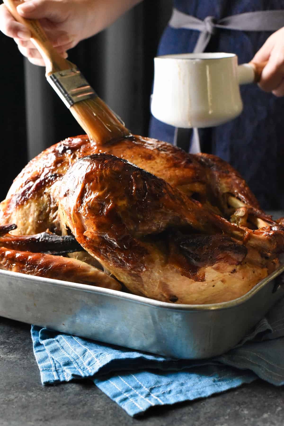 Roasting pan with cooked turkey rests on table. Woman in denim apron is standing behind turkey, glazing it with a basting brush and small white pot of maple bourbon glaze..