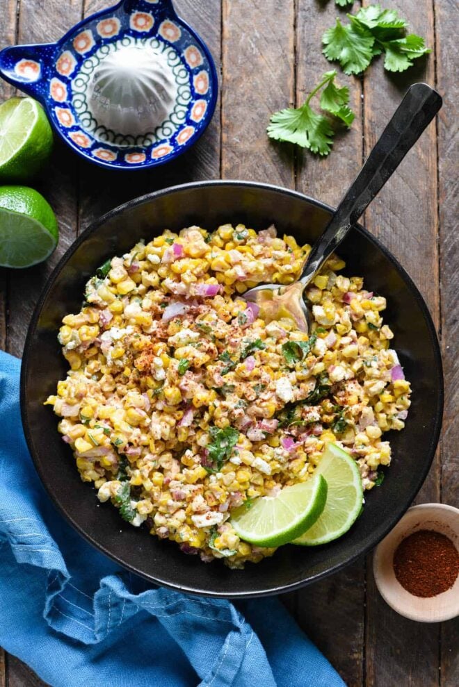 Mexican roasted corn salad in a black bowl, garnished with lime wedges.