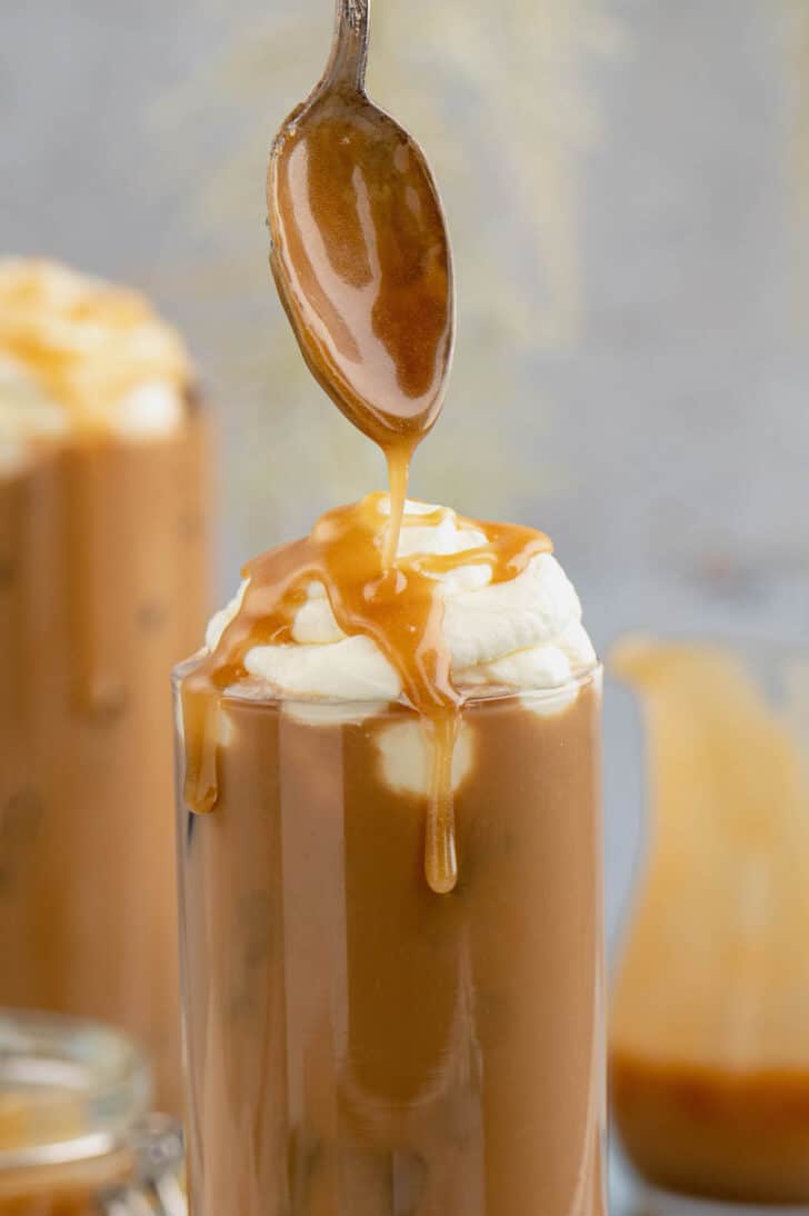 A spoon drizzling a light brown dessert syrup over an iced coffee topped with whipped cream.