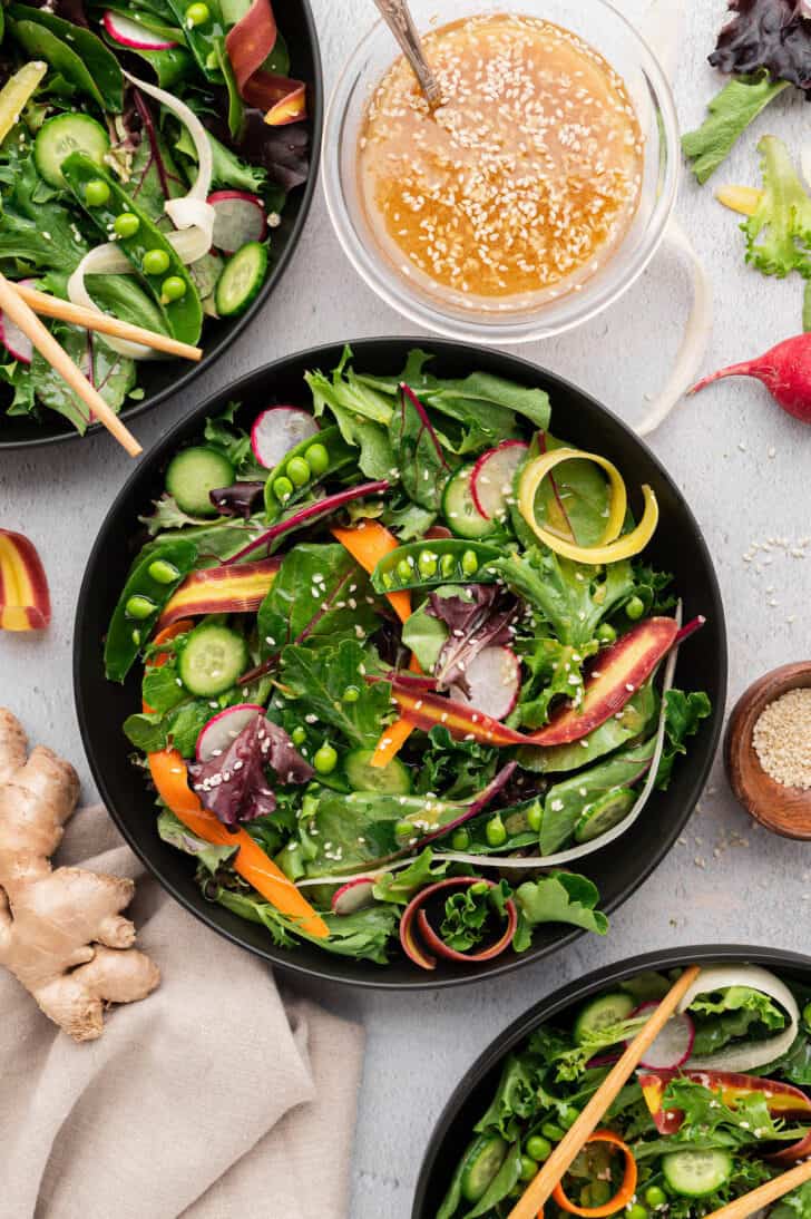 Two shallow black bowls filled with fresh vegetable salad alongside a small glass bowl of miso sesame ginger dressing.