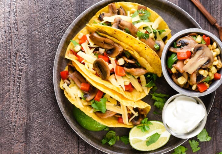 A round tray filled with with three corn tortillas filled with sauteed vegetables, a lime wedge, and small bowls of vegetable filling and sour cream.