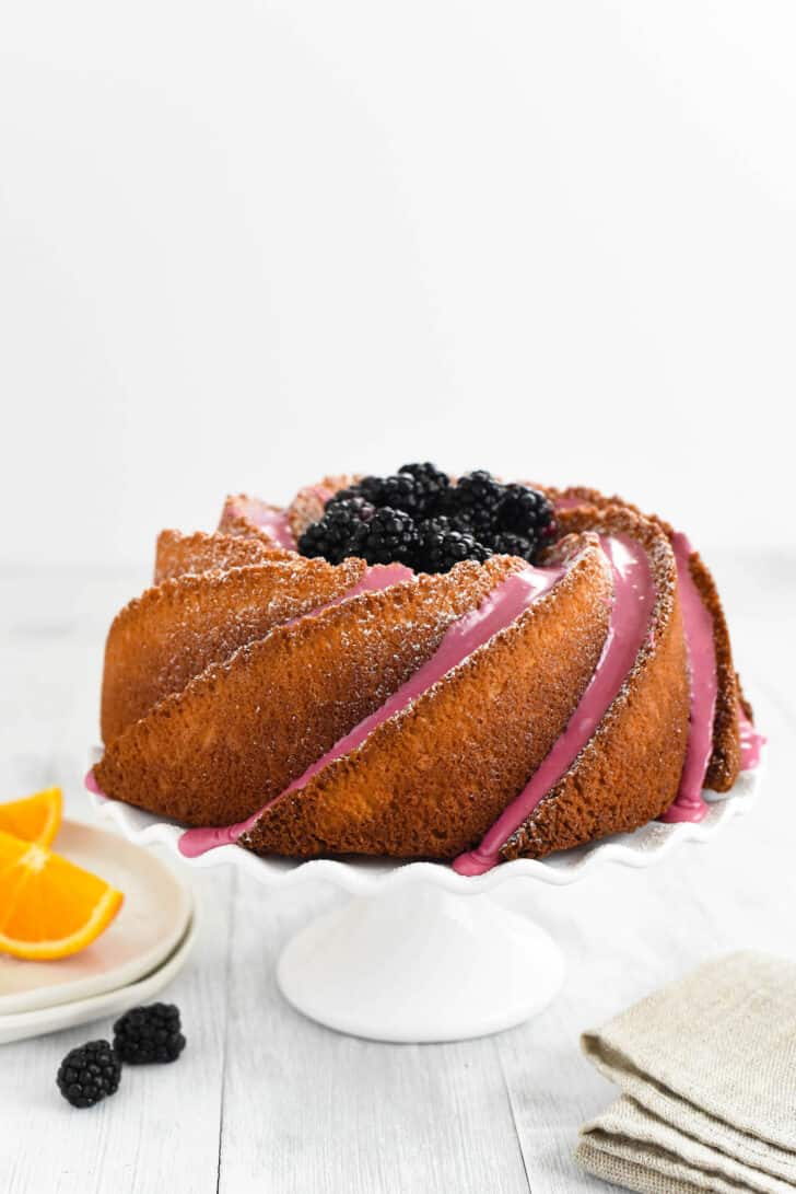 An orange bundt cake decorated with blackberry icing and fresh blackberries, on a white fluted cake stand.