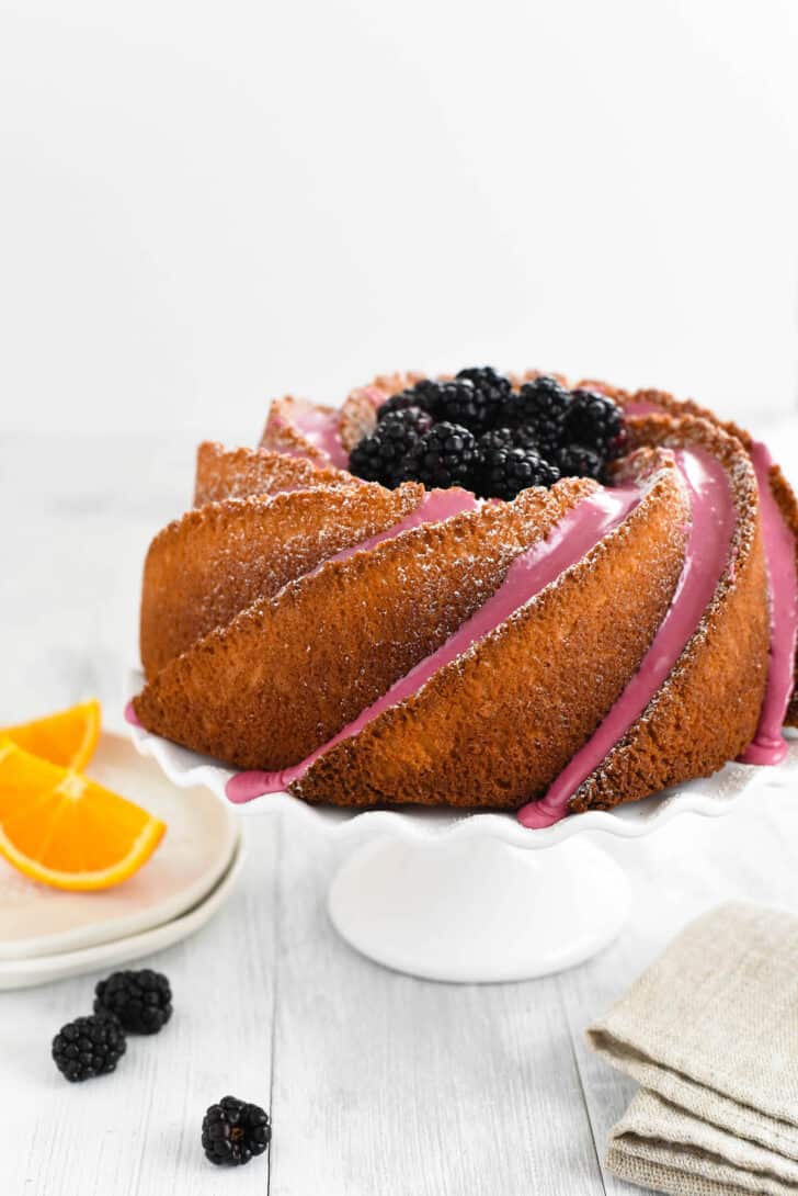 An orange bundt cake decorated with blackberry icing and fresh blackberries, on a white fluted cake stand.