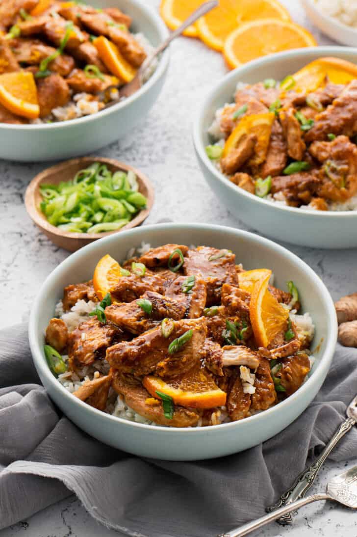 White bowls filled with rice and orange ginger chicken, garnished with orange slices and chopped green onions.