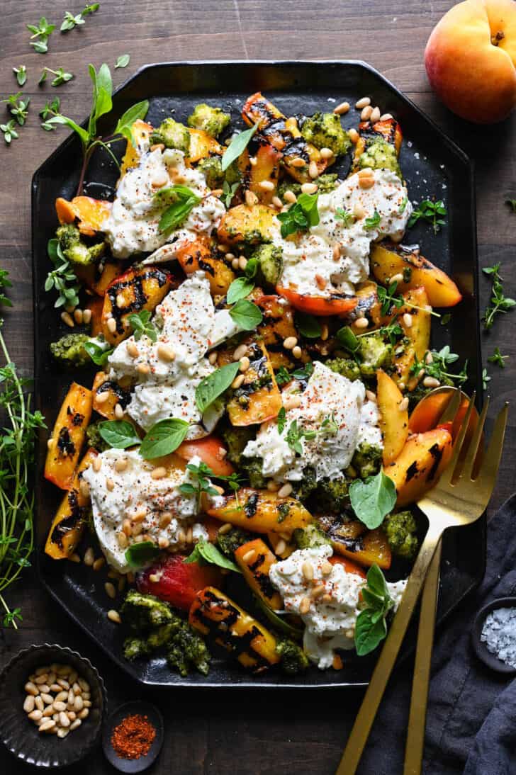 A large black geometric platter piled high with grilled peach burrata salad, garnished with lots of fresh herbs and pine nuts.