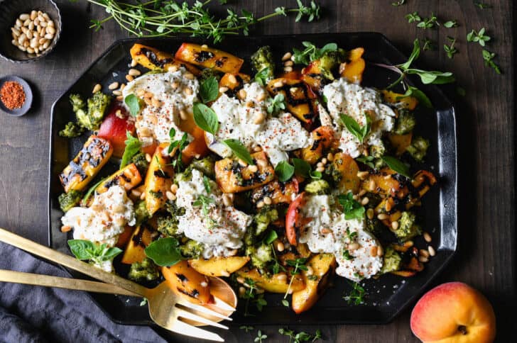 A large black geometric platter piled high with grilled peach salad with burrata, garnished with lots of fresh herbs and pine nuts.