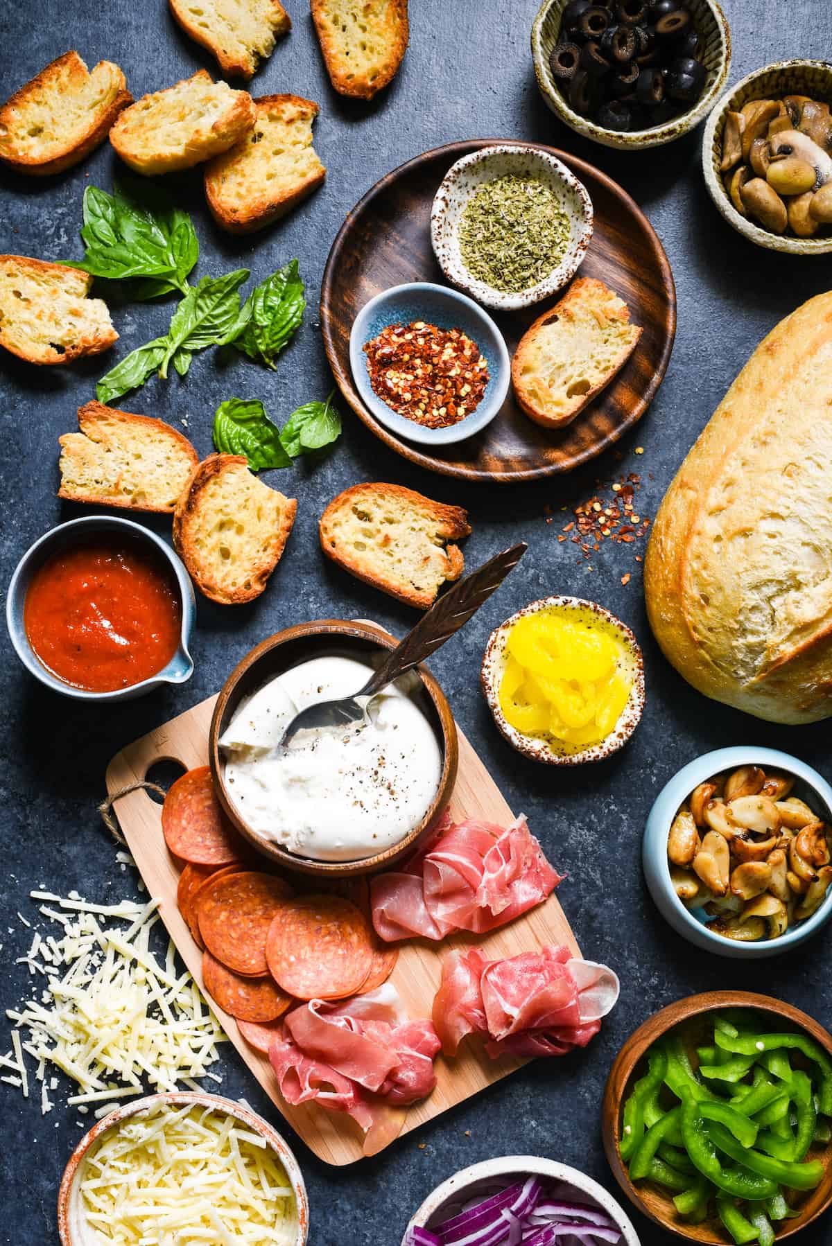 A dark surface filled with the toppings for a crostini pizza bar, including toasted bread, meats, cheese, vegetables and herbs and spices.