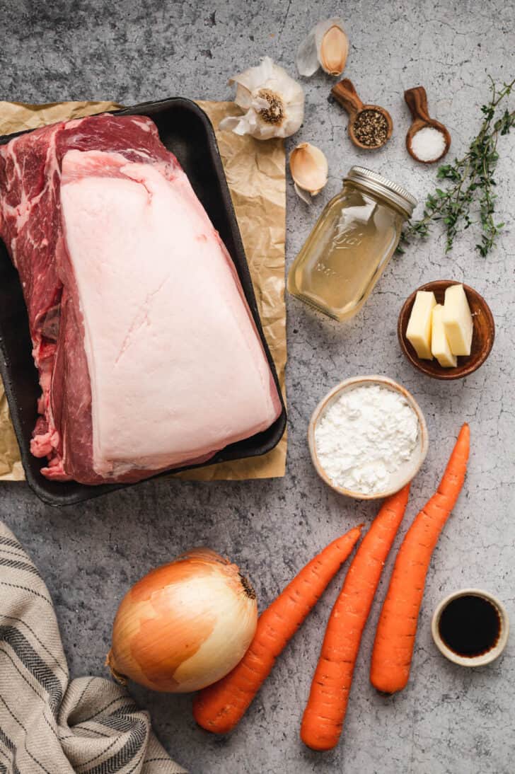 Ingredients laid out on a light surface, including a big piece of meat, carrots, onion, butter, herbs spices, garlic, flour and soy sauce.