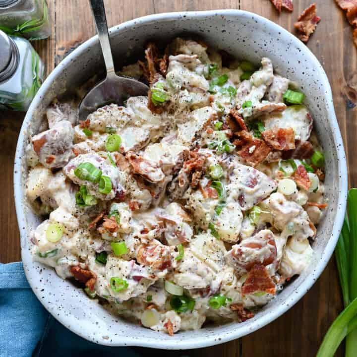 A speckled ceramic bowl filled with potato salad with bacon and green onions, with a spoon digging into the salad.