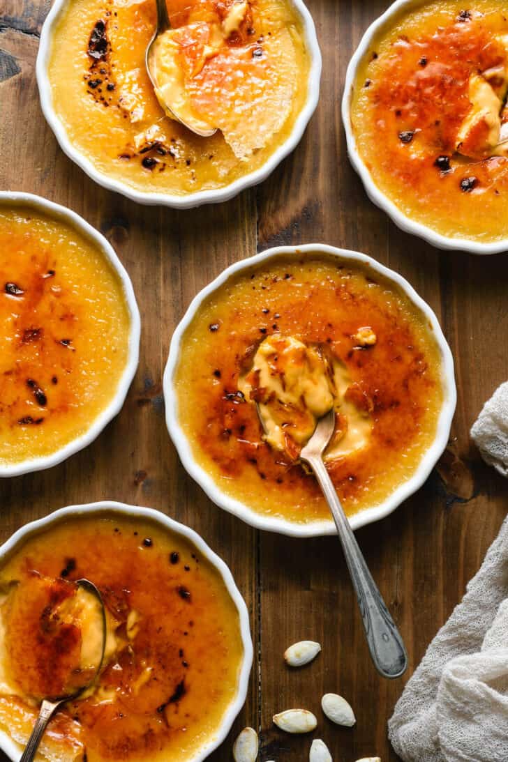 Shallow white ramekins filled with a fall spiced orange-huged custard on a wooden tabletop, with pumpkin seeds and spoons garnishing the scene.