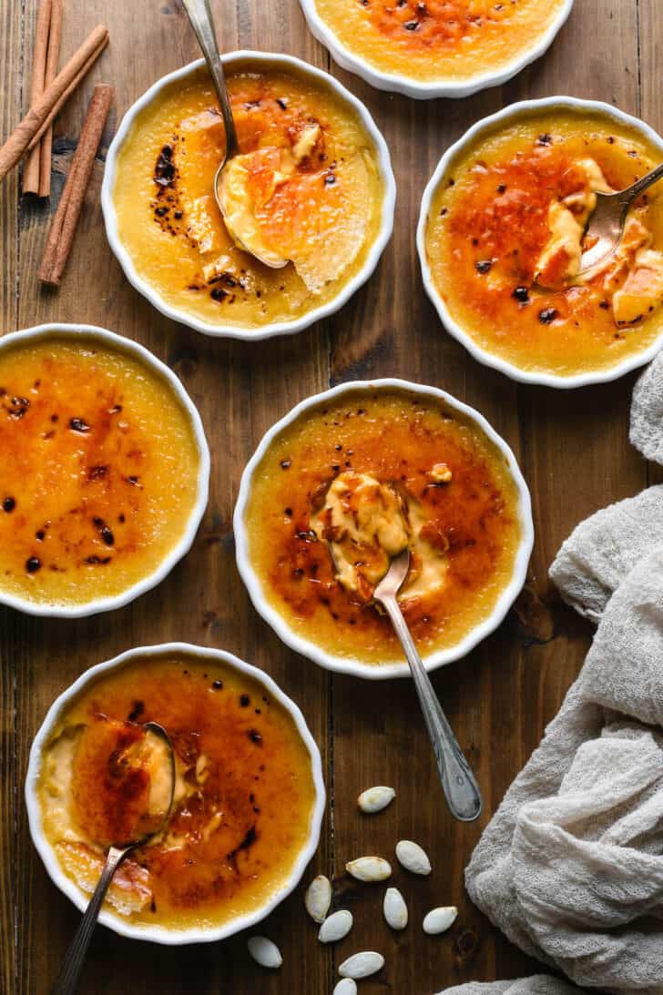 Shallow white ramekins filled with pumpkin creme brulee on a wooden tabletop, with pumpkin seeds, cinnamon sticks, a grey linen and spoons garnishing the scene.