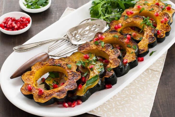 Roasted acorn squash slices garnished with pomegranate seeds and chopped parsley, lined up on a white platter.