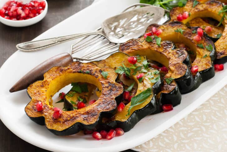 Roasted acorn squash slices garnished with pomegranate seeds and chopped parsley, lined up on a white platter.