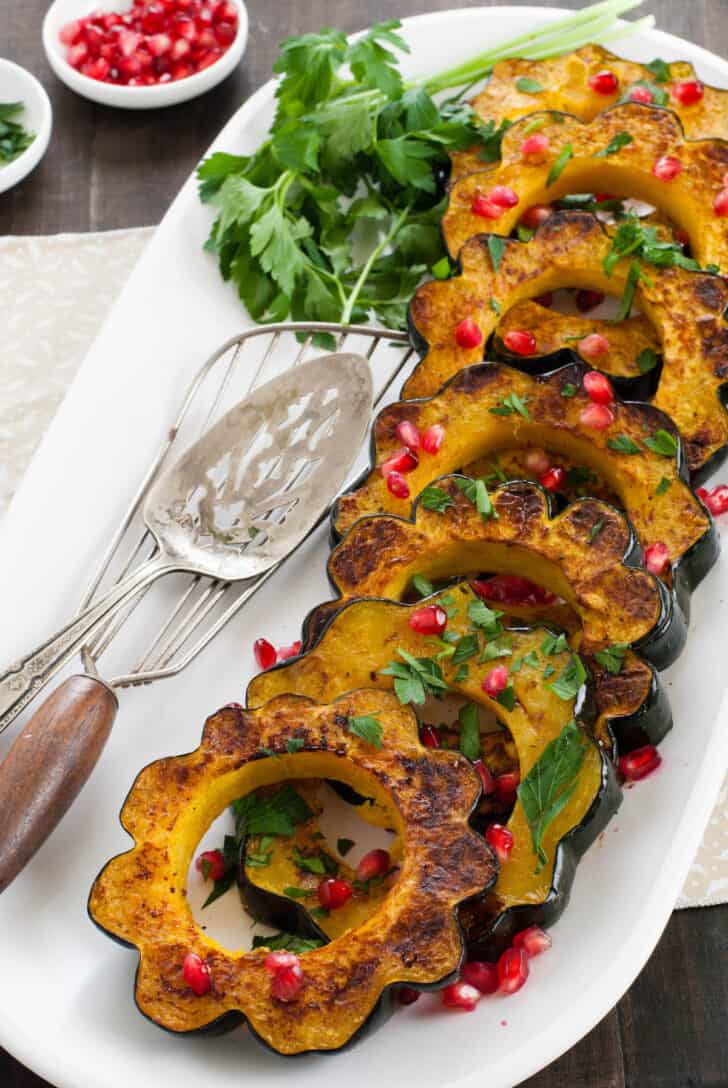 Roasted acorn squash slices garnished with pomegranate seeds and chopped parsley, lined up on a white platter.